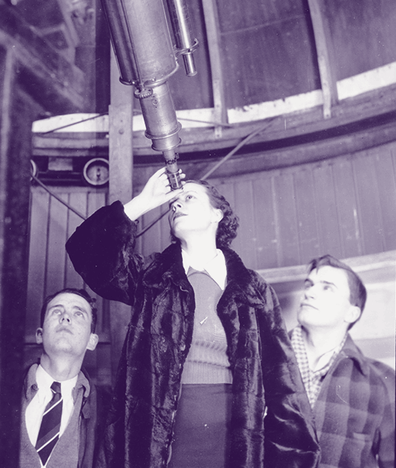 A woman up through a large telescope while two men stand at her side looking into the night sky.